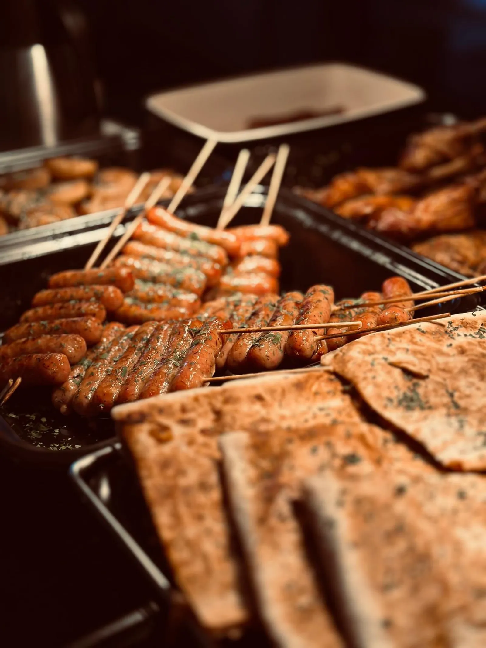 Tarjoiluvuoissa grillattuja makkaroita vartaissa, etualalla yrteillä ripoteltuja lavash-leivän paloja.