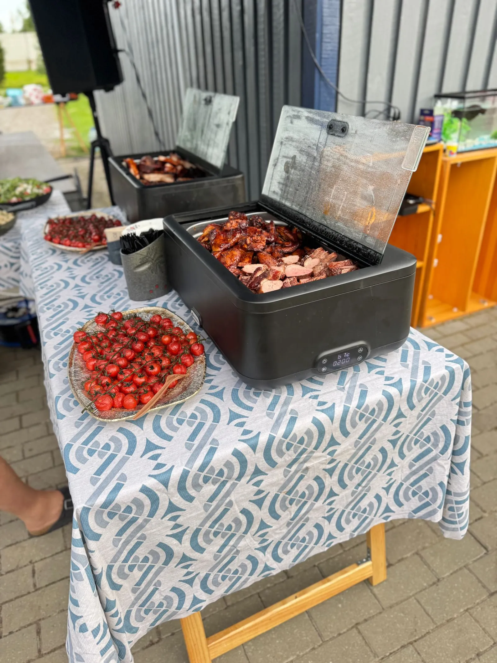 Ulkona katettu pöytä, jossa kaksi avattua lämpölaatikkoa glaseerattuja lihapaloja ja viipaloitua lihaa, vieressä korillinen kirsikkatomaatteja ja lautasellinen punaisia marjoja.
